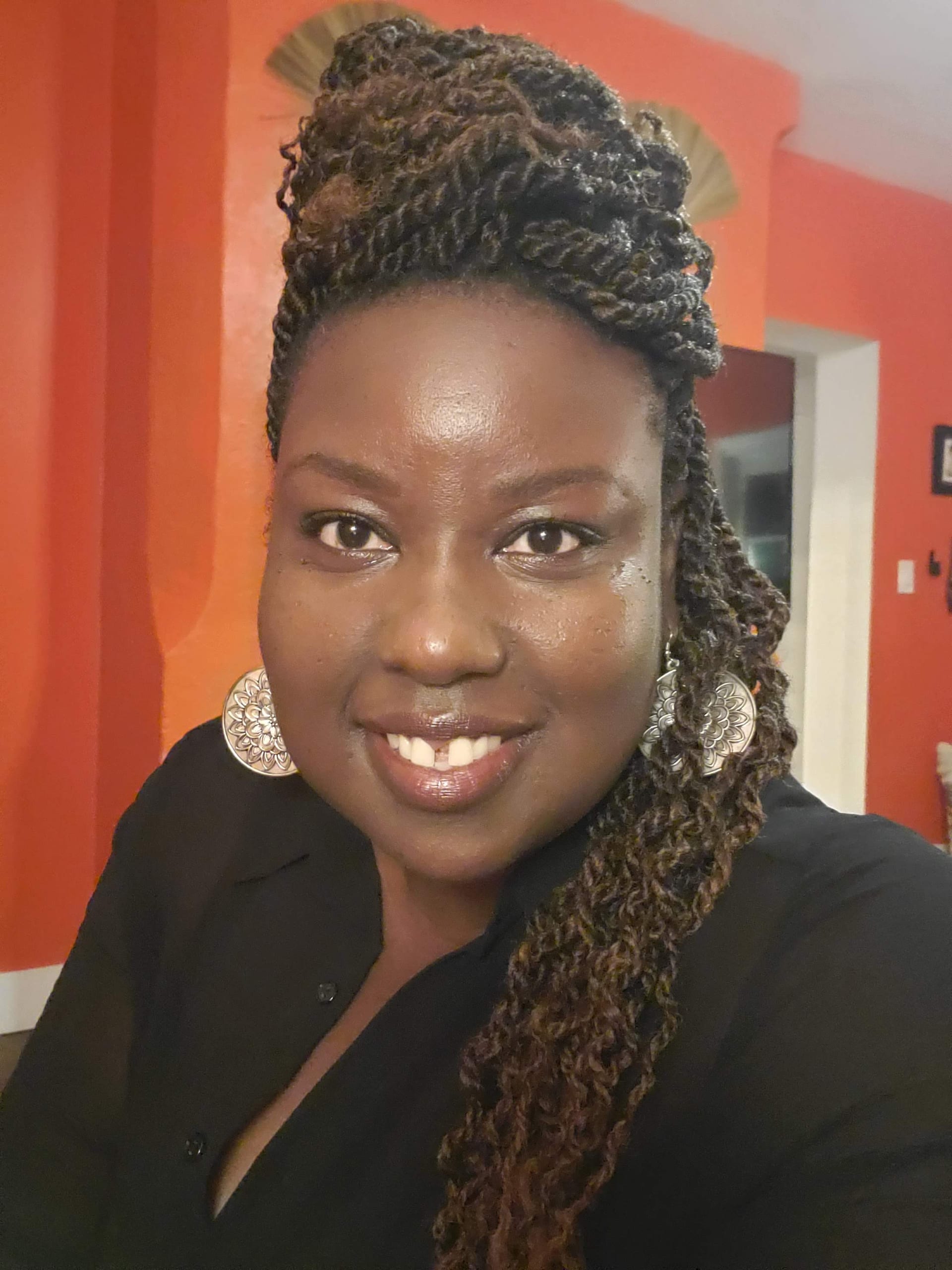 Kali Shelton, close-up portrait with braids in a black shirt against a warm wall